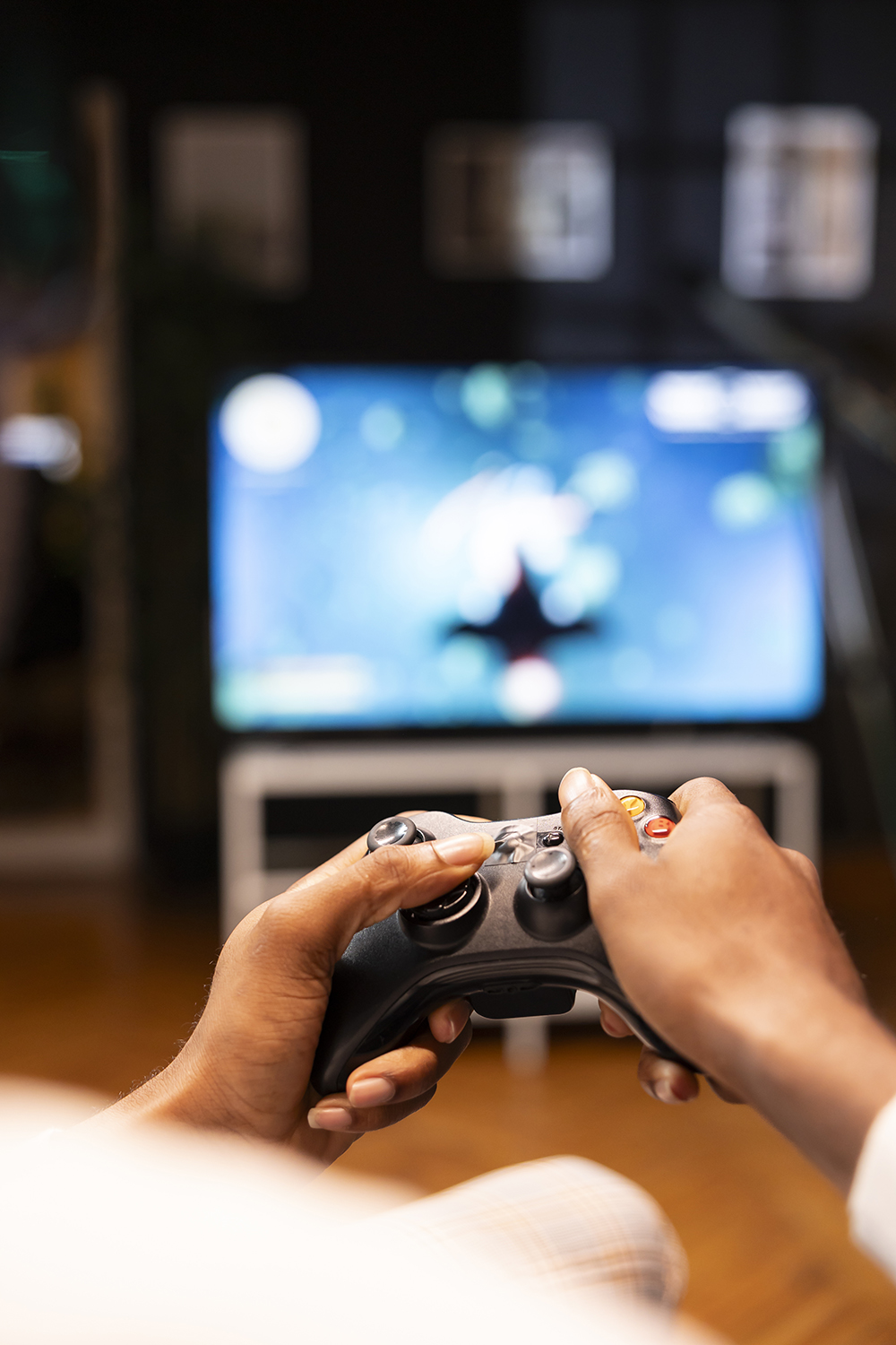 Focus shot on man holding controller in apartment, playing videogames on smart TV display in blurry background. Gamer on sofa using joypad to participate in game on console attached to television set