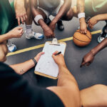 Basketball, strategy and team with a sports coach talking to a team while planning tactics on a clipboard and hands. Teamwork, fitness and exercise with a player and teammates listening at training.