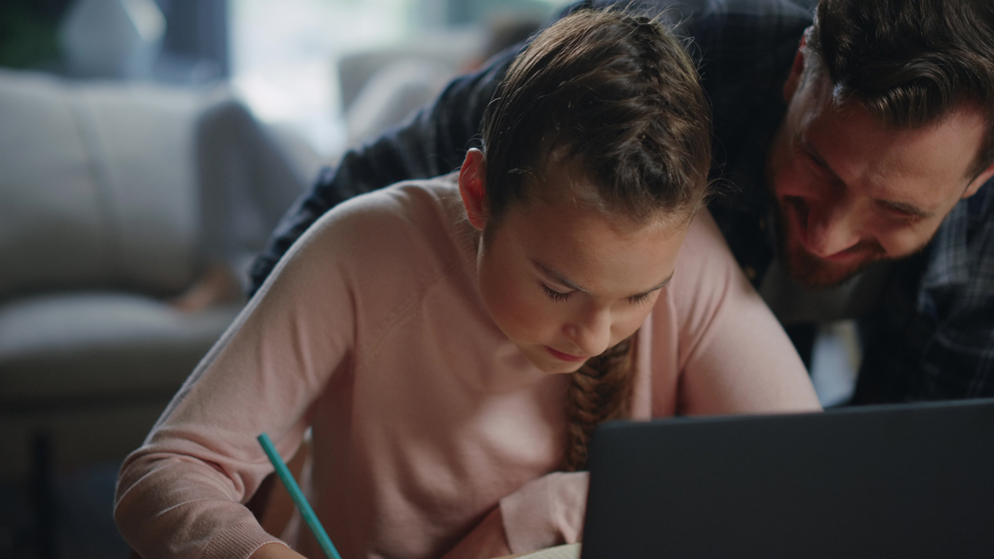 Closeup smiling father helping daughter with homework at table with laptop. Teen girl studying remote under parent supervision at home. Diligent child writing notebook next to father indoors.