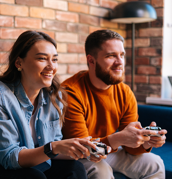 Happy cheerful young couple holding controllers and playing video games on console sitting together on couch at cozy living room. Concept of leisure activity of lovers at home.